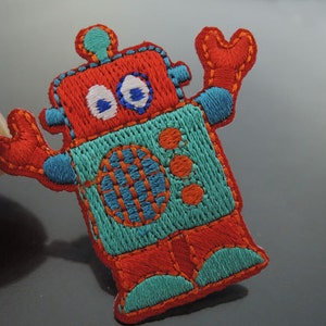 Könnte beinhalten: Gestickter Roboter-Patch in Rot und Blau mit weißen Augen. Der Roboter hat einen großen, runden Kopf und zwei Arme, die in die Luft gestreckt sind.