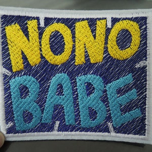 Può includere: Una toppa bianca con tessuto blu al centro. Le parole "NONO BABE" sono ricamate in filo giallo e blu.