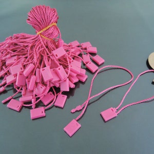 May include: A pile of pink string and plastic tags. The tags are square shaped with a hole in the center. The string is tied together with a rubber band.