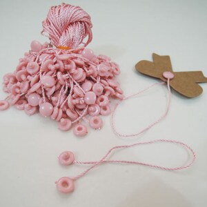 May include: A pile of pink plastic string tags with a brown paper tag with a bow on the side. The string tags have a loop at the top and a small button at the bottom.
