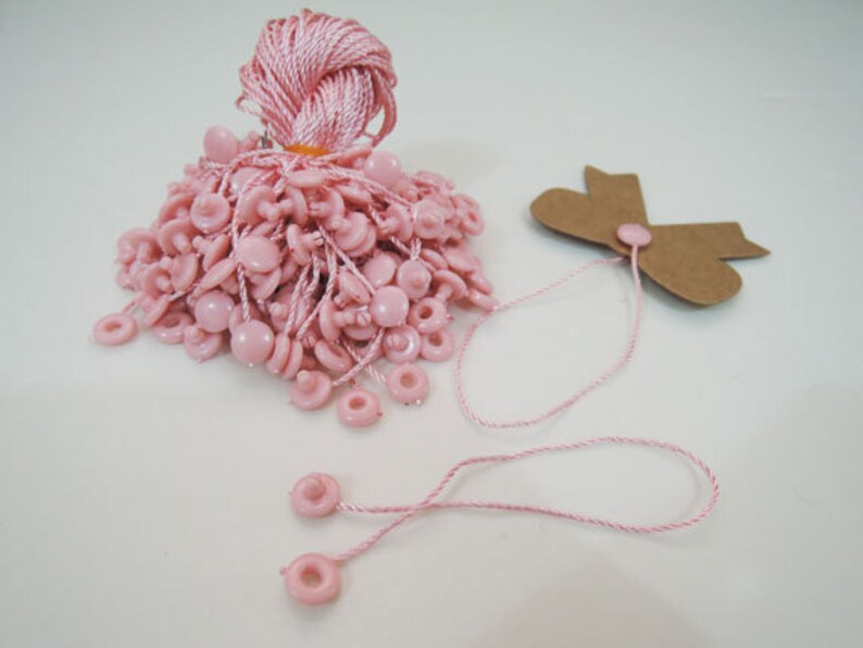 May include: A pile of pink plastic string tags with a brown paper tag with a bow on the side. The string tags have a loop at the top and a small button at the bottom.
