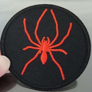 Könnte beinhalten: Ein schwarzer runder Patch mit einem roten gestickten Spinnenmotiv.