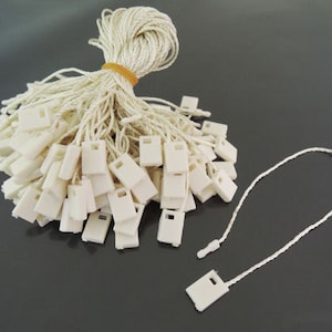 May include: A pile of white string tags with a small white plastic loop at the end of each string. The tags are bundled together with a rubber band.
