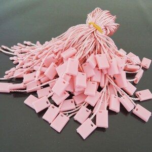 May include: A bundle of pink rectangular price tags with white strings. The tags are made of plastic and have a small hole at the top. The strings are tied together with a pink cord and secured with a yellow rubber band.