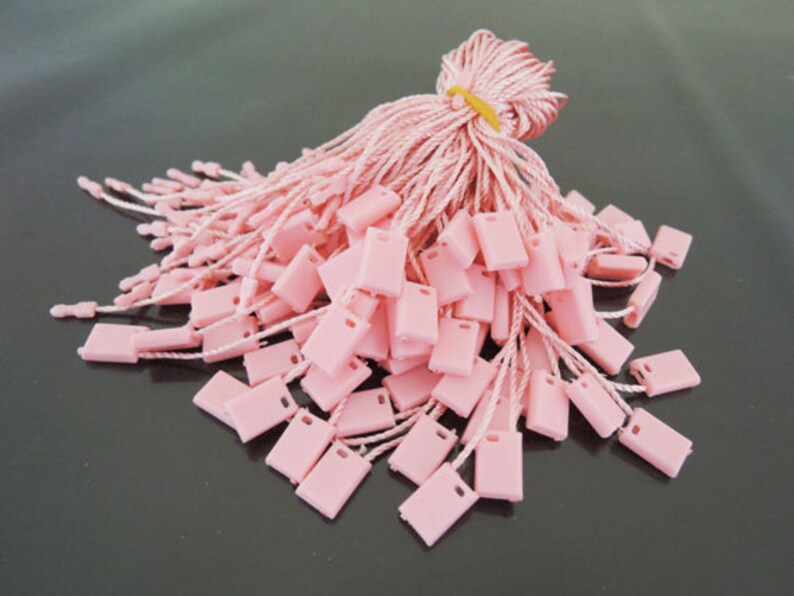 May include: A bundle of pink rectangular price tags with white strings. The tags are made of plastic and have a small hole at the top. The strings are tied together with a pink cord and secured with a yellow rubber band.