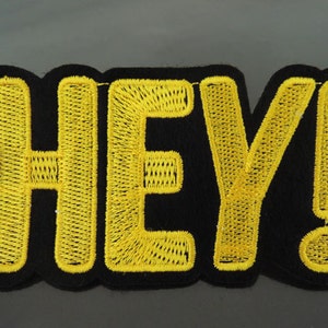 May include: Embroidered patch with the word "HEY!" in bright yellow thread on a black background. The patch has a scalloped edge and a bold, eye-catching design. The exclamation point adds emphasis to the message.