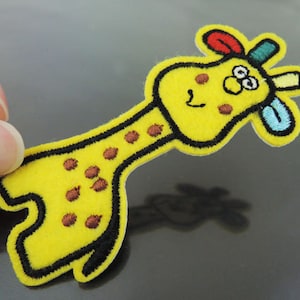 Op de afbeelding: Gele vilt giraffe applicatie met bruine vlekken, zwarte omtrek en kleurrijke details op het hoofd.