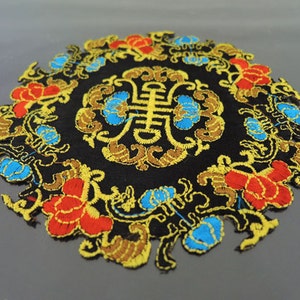 Könnte beinhalten: Ein runder gestickter Aufnäher mit schwarzem Hintergrund und aufwendigen goldenen, roten und blauen Blumendesigns. Die Mitte des Aufnähers zeigt ein stilisiertes chinesisches Schriftzeichen.