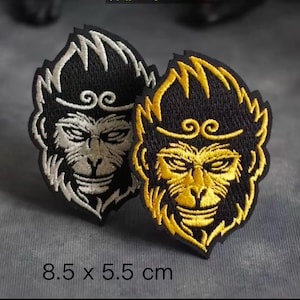 Op de afbeelding: Twee geborduurde strijkpatches met een apenkop met een gouden halo. De aap links is wit en de aap rechts is goud. Beide patches zijn 8,5 x 5,5 cm.