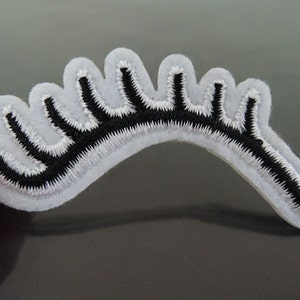 Pode incluir: Aplique de cílios de feltro branco com bordado preto. O cílio é curvo e tem uma borda festoneada.