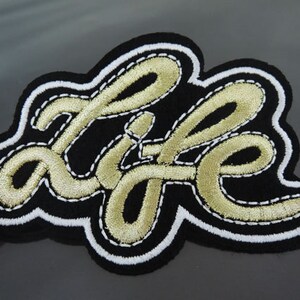 May include: Embroidered patch with the word "LIFE" in gold cursive lettering on a black background. The patch has a white border with gold stitching, creating a decorative and eye-catching design.