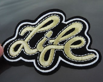 May include: Embroidered patch with the word "LIFE" in gold cursive lettering on a black background. The patch has a white border with gold stitching, creating a decorative and eye-catching design.