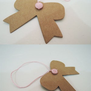 May include: A brown paper bow with a pink button in the center. The bow is attached to a pink and white string.