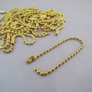 May include: A close-up of a gold ball chain with a small connector. The chain is made of small, round beads and is lying on a light purple surface.