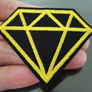 Könnte beinhalten: Schwarzer Diamant-förmiger Patch mit gelber Umrandung. Der Patch ist aus Stoff und hat ein gesticktes Design.