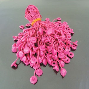 May include: A pile of pink string tags with a plastic button at the end of each string. The strings are bundled together with a yellow rubber band.