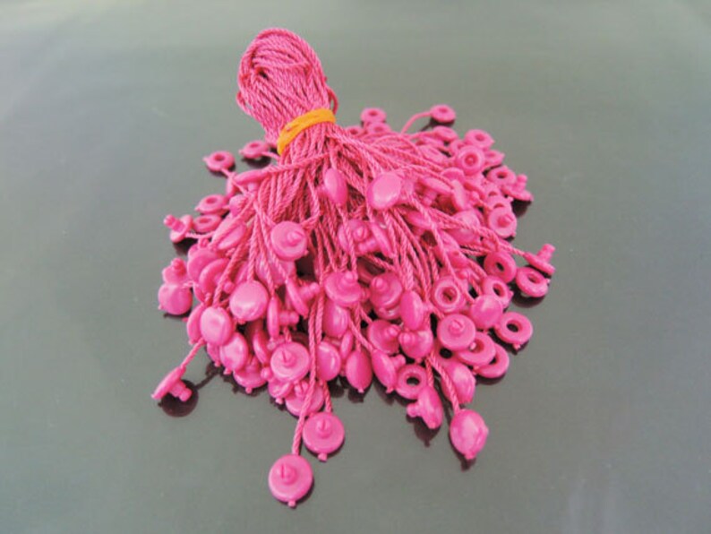 May include: A pile of pink string tags with a plastic button at the end of each string. The strings are bundled together with a yellow rubber band.