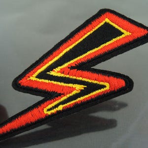 May include: A red and yellow lightning bolt shaped patch with black stitching. The patch is made of felt and has a black backing.