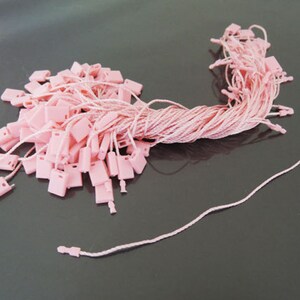 May include: A pile of pink hang tags with matching string. The tags are rectangular with a small hole at the top for the string. The string is a light pink color and is attached to each tag.