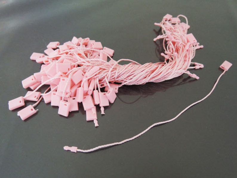 May include: A pile of pink hang tags with matching string. The tags are rectangular with a small hole at the top for the string. The string is a light pink color and is attached to each tag.