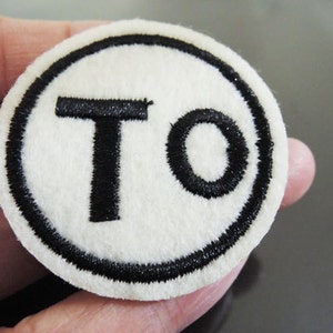 Könnte beinhalten: Weißer Filz-Kreis-Patch mit schwarzem gesticktem Rand und den Buchstaben "TO" in Schwarz.