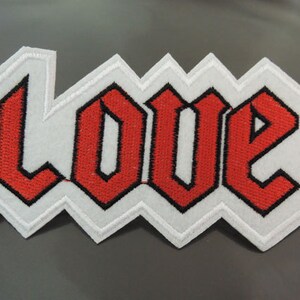 Könnte beinhalten: Roter gestickter Aufnäher mit dem Wort "LOVE" in gotischer Schrift. Der Aufnäher ist weiß mit schwarzen Umrandungen um die Buchstaben.