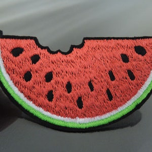 以下が含まれることがあります： 一口かじられた赤いスイカの切り身。スイカは黒い種が刺繍されており、白と緑の縁取りがあります。