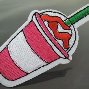 Pode incluir: Etiqueta bordada rosa e branca de um milkshake com um canudo verde.