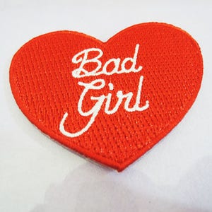 May include: Red heart-shaped patch with white embroidered text that reads "Bad Girl".