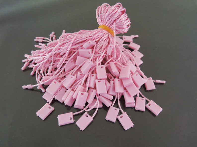 May include: A pile of pink string tags with square plastic ends. The tags are bundled together with a rubber band.