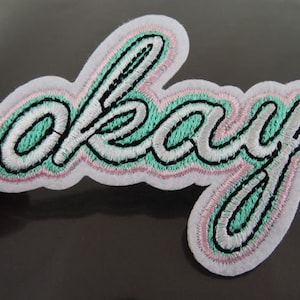 May include: A white, green, and pink embroidered iron-on patch with the word "okay" in a cursive font.