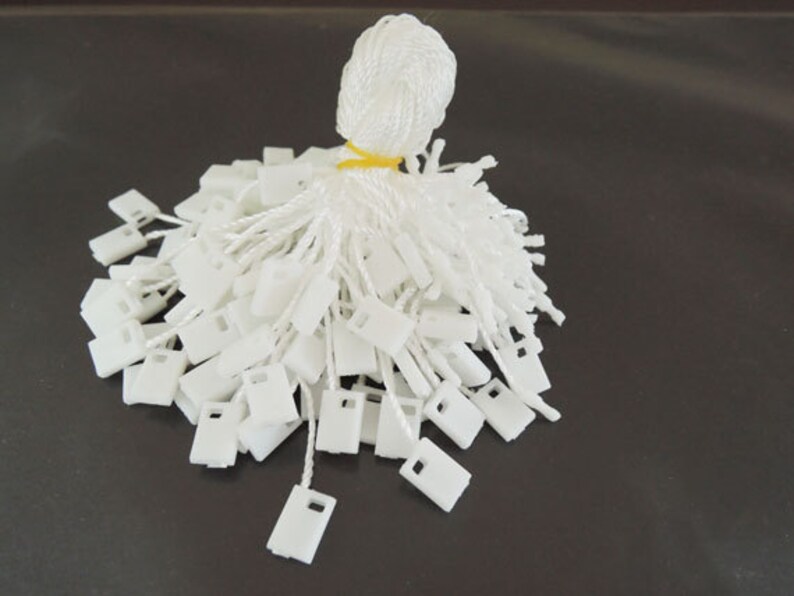 May include: A pile of white plastic string tags with a white string attached to each tag. The tags are arranged in a pile with the strings sticking out.