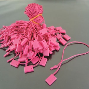 May include: A pile of pink string tags with a square loop at the end. The tags are tied together with a rubber band.