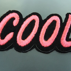 Könnte beinhalten: Ein schwarzer Filz-Patch mit dem Wort "COOL" in rosa Stickerei.