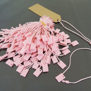 May include: A collection of pink rectangular price tags with attached strings, bundled together with a brown tag and a yellow rubber band. The tags are scattered on a dark surface, with some strings extending outwards.