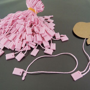 May include: A pile of pink string with small square plastic tags attached. The string is tied together with a yellow rubber band. A single string with a tag is laid out on a black surface.