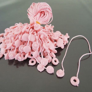 May include: A pile of pink string tags with a small round button at the end of each string. The tags are bundled together and there is a loose string tag with a button at the end.