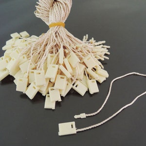 以下が含まれることがあります： ベージュのプラスチック製のタグが紐で結ばれています。タグは正方形で、上部に小さな穴が開いています。紐はベージュで、束ねられています。