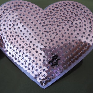 May include: A pink heart-shaped sequin patch. The sequins are arranged in a circular pattern and have a metallic sheen.