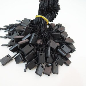 May include: A pile of black plastic tags with black string attached. The tags are square shaped and have a small hole in the center.