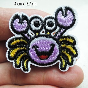 Könnte beinhalten: Lila und gelbe gestickte Bügelapplikation mit einem lächelnden Cartoon-Krabben. Die Krabbe hat große Augen und einen fröhlichen Ausdruck. Die Applikation misst 4 cm x 3,7 cm.