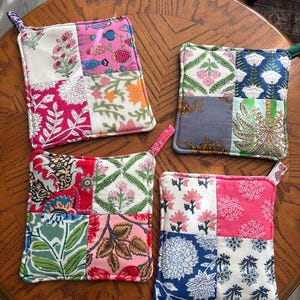 May include: Four square quilted pot holders featuring patchwork designs with various floral, fish, and animal patterns in vibrant colors including pink, blue, green, and white. Each piece has a small fabric loop attached to one corner for hanging.