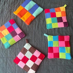 May include: Four square fabric potholders featuring patchwork designs in various bright colors including red, orange, yellow, green, blue, purple, pink, and brown. Each potholder has a small fabric loop attached to one corner for hanging purposes.