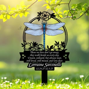 May include: Black metal memorial stake featuring a dragonfly design with light blue wings and a blue body. The stake includes a circular frame with floral accents and a banner with the text "Those we love don't go away..." and the name "Lorraine Sarcinelli".