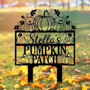 Puede incluir: Letrero de metal negro con el texto "Stella's PUMPKIN PATCH" y diseños de calabazas y hojas. El letrero está clavado en el suelo, ideal para un jardín o patio. El letrero está rodeado de hojas caídas.