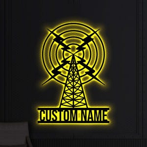 May include: Black metal wall art depicting a radio tower with a yellow illuminated outline. The design includes concentric circles, lightning bolts, and the text "CUSTOM NAME" in a rectangular base. The art is mounted on a textured black wall.