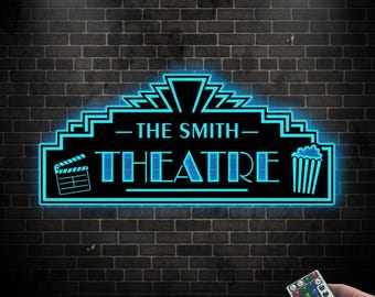 May include: A black and blue neon sign for "THE SMITH THEATRE" with a film clapboard and popcorn bucket icon. The sign is mounted on a dark brick wall. A hand holds a remote control with colorful buttons.