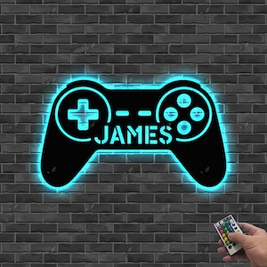 May include: A black, illuminated gaming controller-shaped sign with the name "JAMES" in white. The sign has a blue neon glow and is mounted on a dark gray brick wall. A remote control is held in the hand.