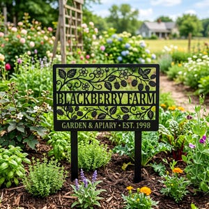 May include: A black metal garden sign with a rectangular frame featuring laser-cut blackberry vines, leaves, and flowers. The text reads BLACKBERRY FARM and GARDEN & APIARY • EST. 1998. The sign is supported by two vertical metal stakes.