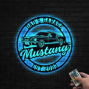 May include: A black metal sign with a blue neon glow, featuring a classic Mustang car silhouette inside a circular frame. The text "DAD'S GARAGE" arches above the car, with "Mustang" in cursive below. The sign includes "EST 2023".